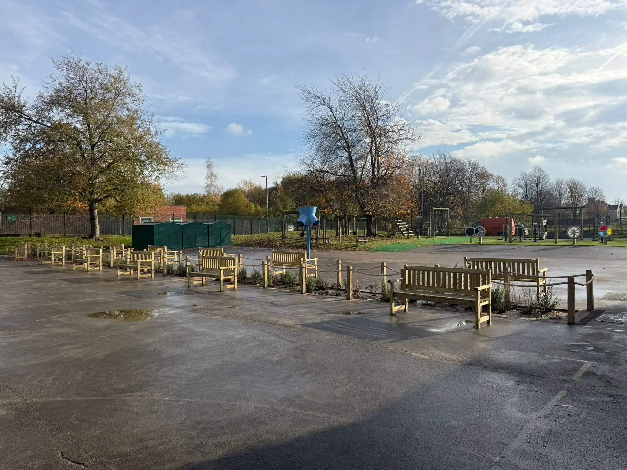 A view of a park with trees, a gravel path, and a cloudy sky in the background. Waterlogged ground visible.