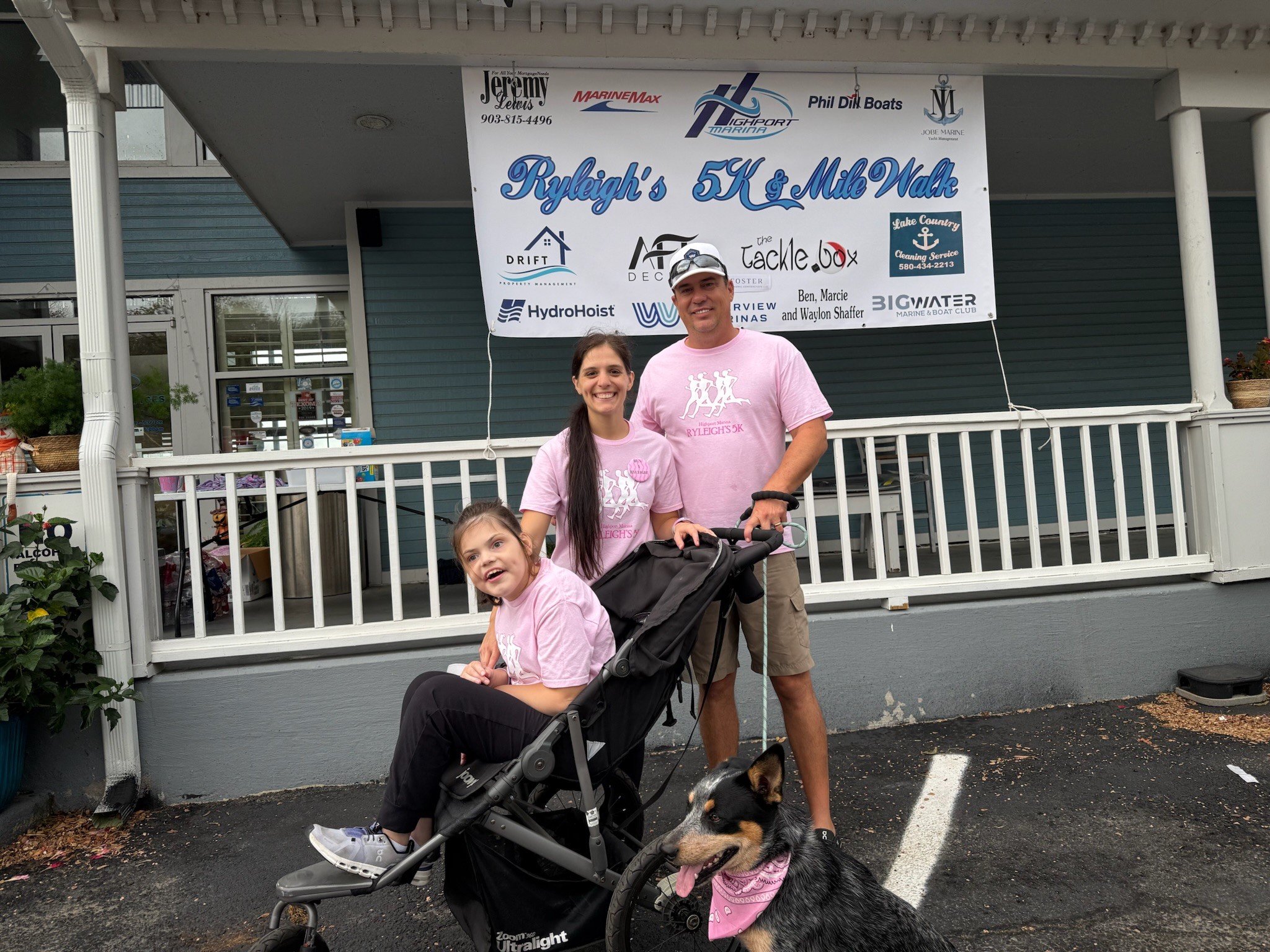 A family stands smiling in front of a building, with a young child in a stroller and a dog wearing a pink bandana, participating in a community fundraising event, as displayed by the banner in the background.