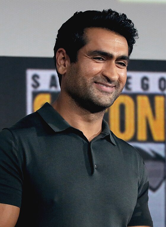 Kumail Nanjiani wearing a dark polo shirt. He stands in front of a blurred background featuring a colourful logo.