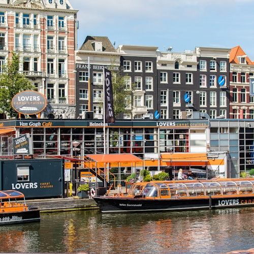 Canal with glass-roof boats labeled "LOVERS" in front of a cafe and colorful buildings lining the street in Amsterdam.