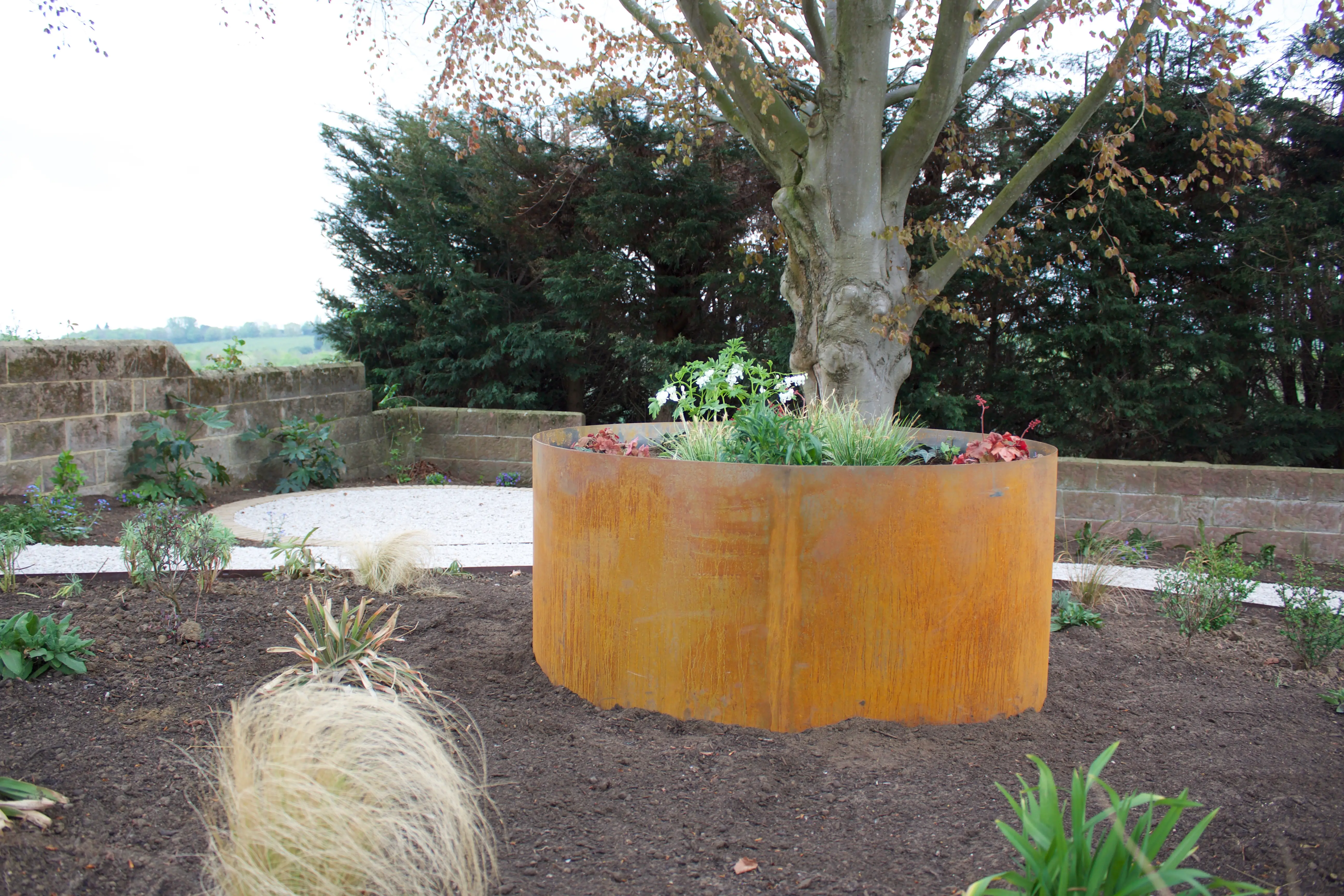 A wooden garden planter filled with greenery, placed outdoors near a tree and surrounded by mulch.