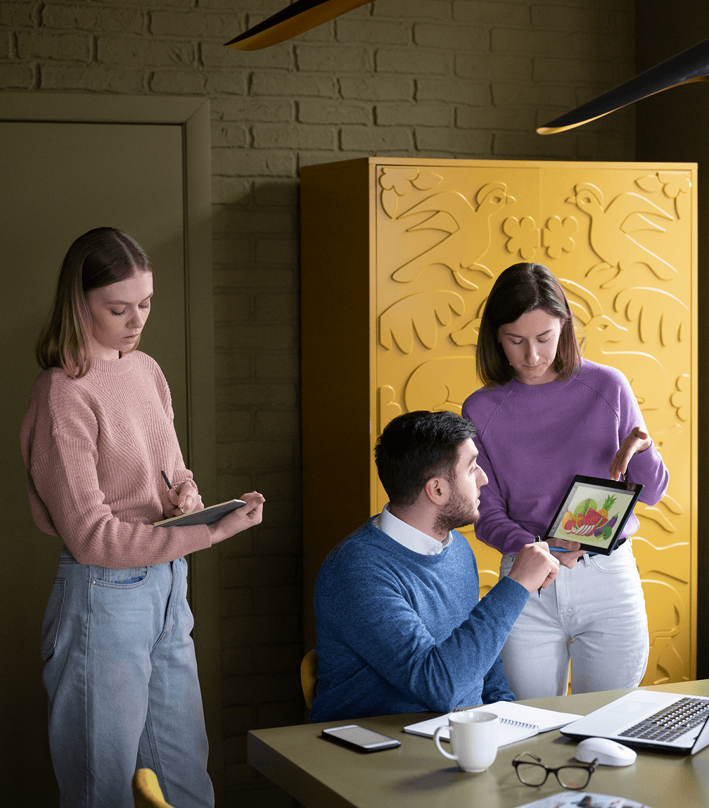 Three individuals gathered around a table, engaged in discussion while examining a laptop.