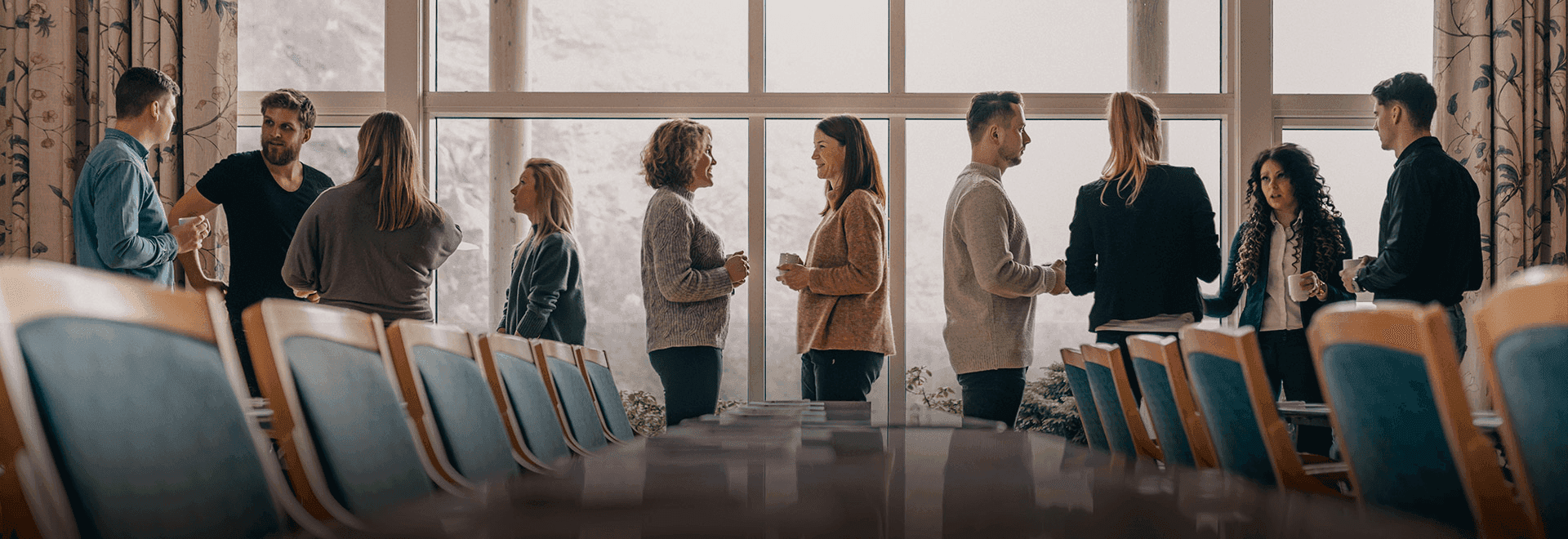 A group of people engaged in conversation stands in a spacious conference room filled with natural light, featuring large windows and chairs surrounding a polished wooden table, highlighting a professional networking event or meeting.