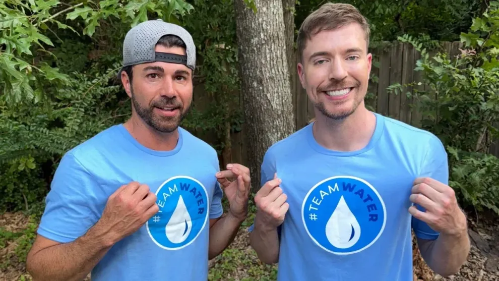 Two men wearing matching blue t-shirts with a water droplet logo pose together in a natural setting, smiling.