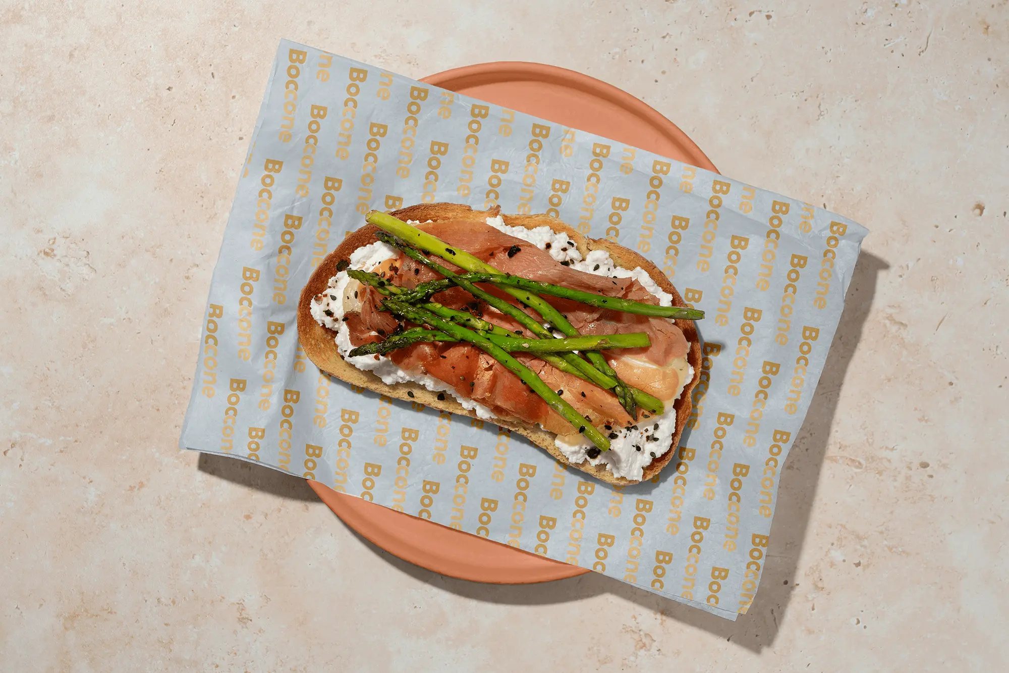 Open-faced Boccone Café toast topped with ricotta, prosciutto, and grilled asparagus served on branded parchment paper over a peach-colored plate.