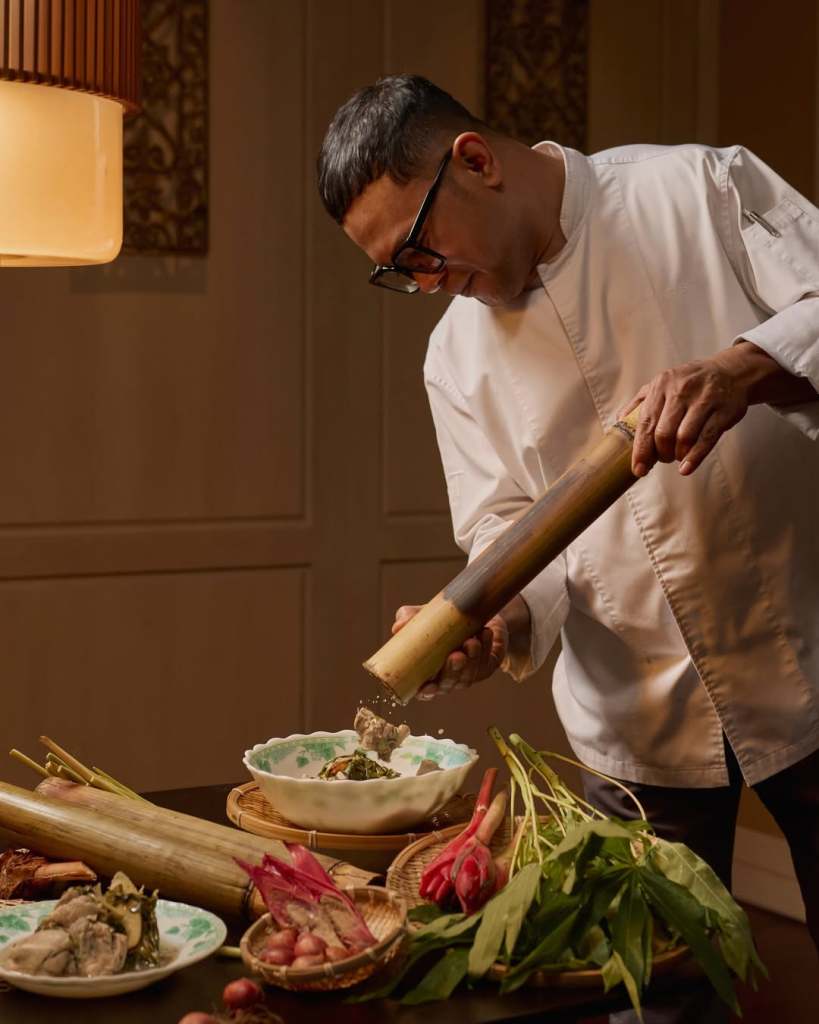 A chef in a white coat uses a bamboo tube to season a dish in a beautifully arranged kitchen setting with fresh ingredients.
