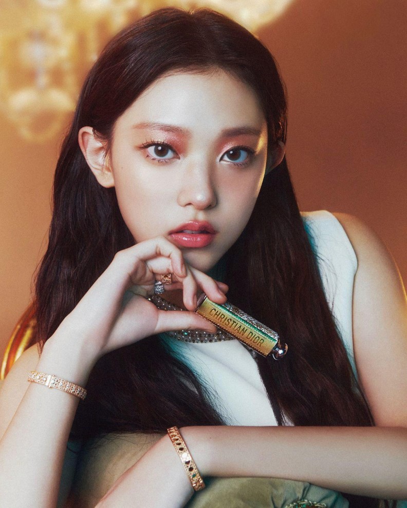 Harin of NewJeans posing with long dark hair, holding a Dior lipstick close to her lips, wearing soft glam makeup and jewelry for Dior Beauty