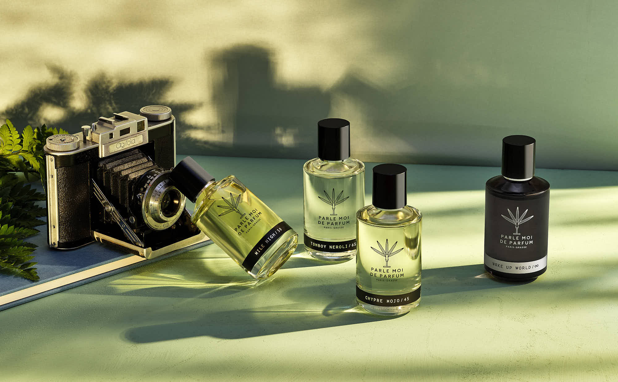 Vintage camera with Parle Moi de Parfum bottles on a green surface, natural light shadows.