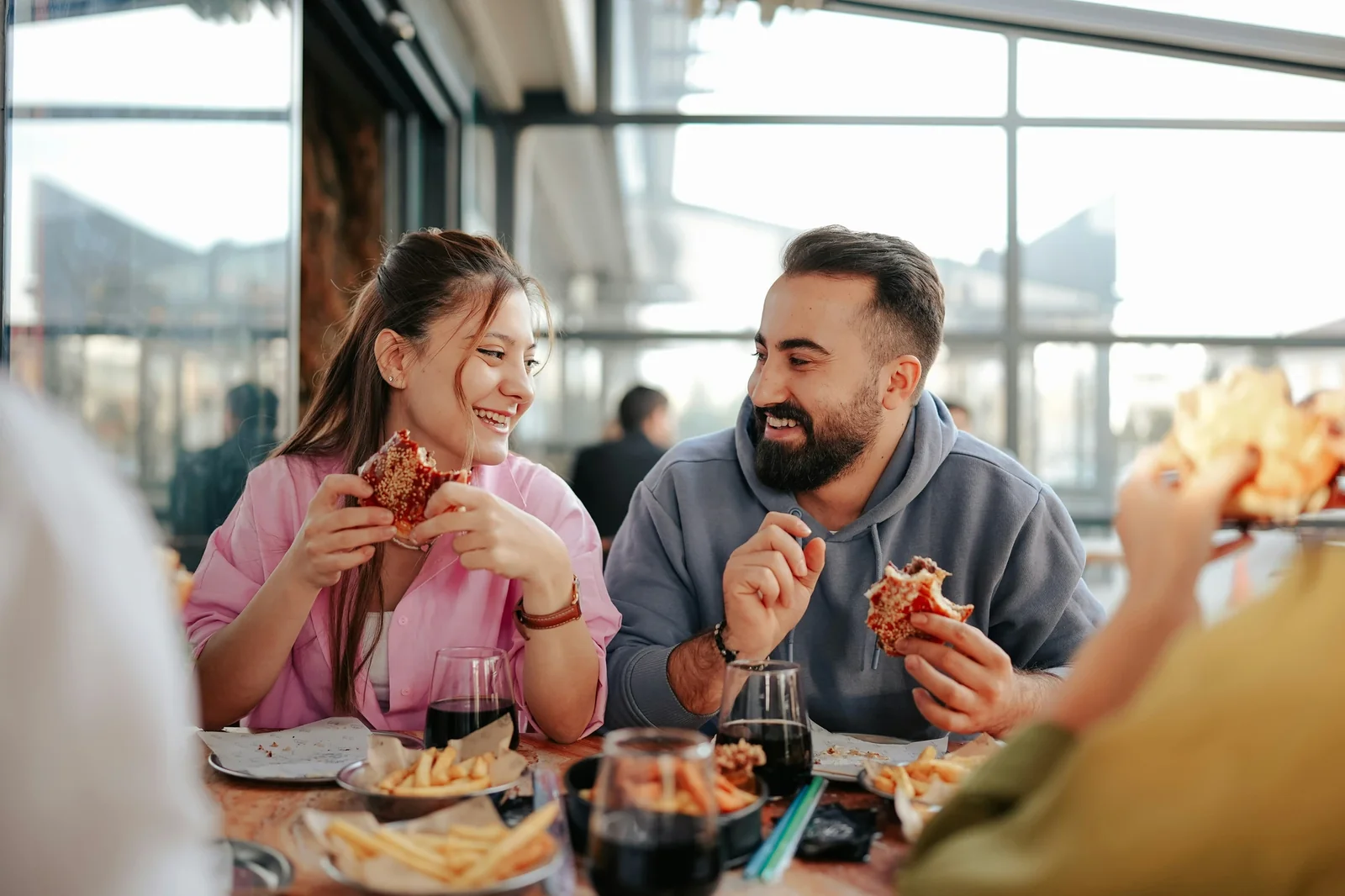 Young couple enjoying burgers and fries at a rooftop restaurant with city skyline view, smiling while eating together.