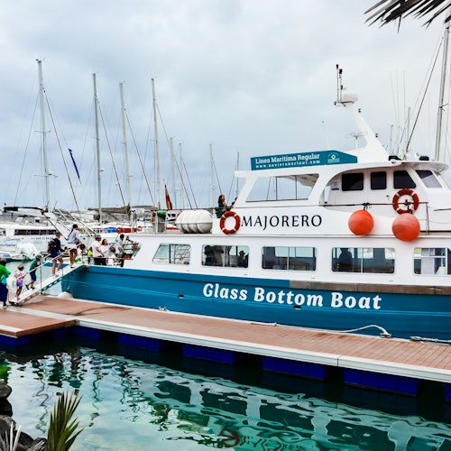 A blue and white glass-bottom boat named 'Majorero' docked at a marina, with people boarding from a wooden pier.