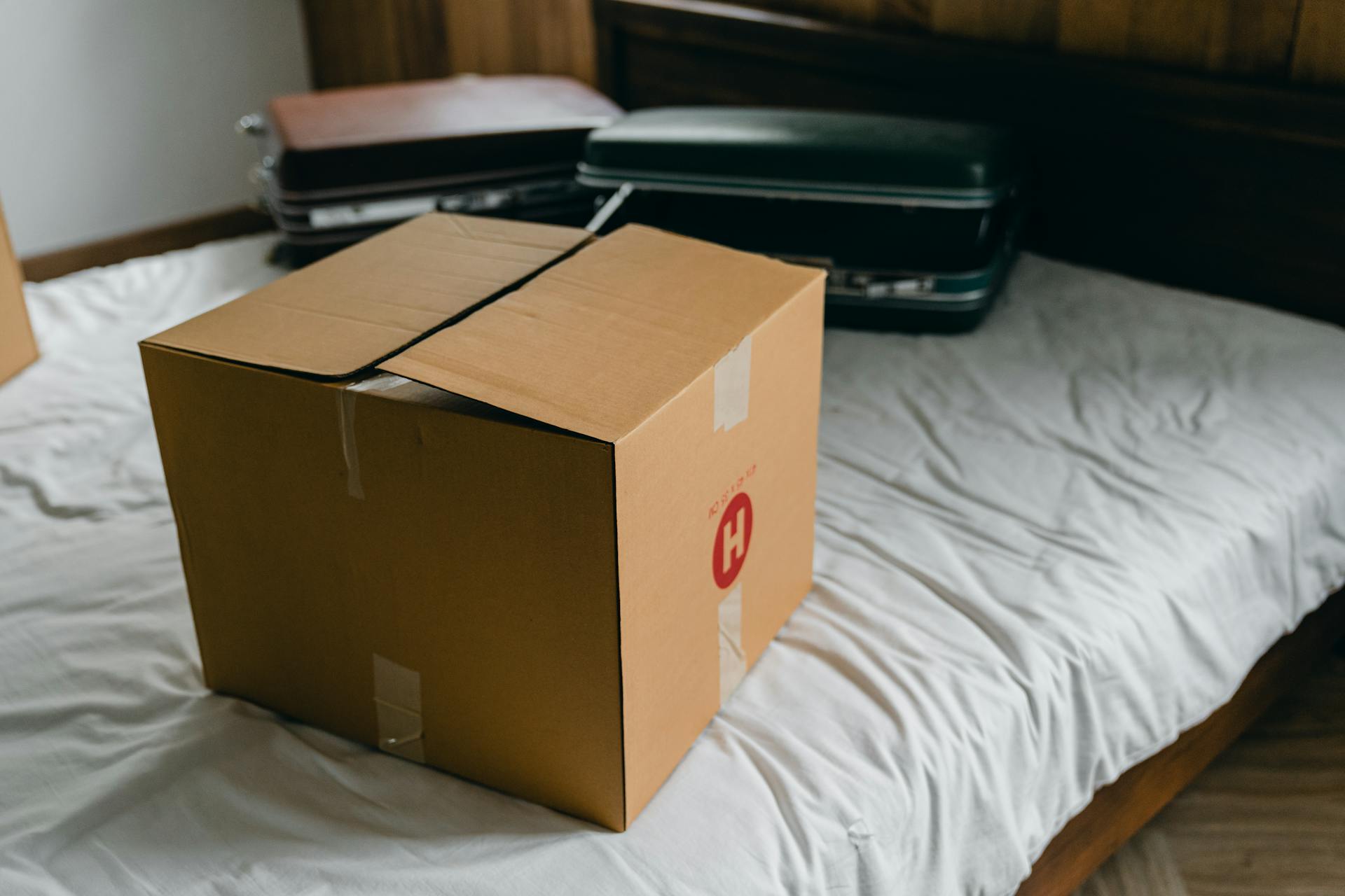 A sealed, medium-sized, plain brown cardboard moving box rests on a white, wrinkled bedspread. The box has a small red logo featuring the letter 'H' on one side. Two vintage-looking suitcases are visible on the bed behind the box.