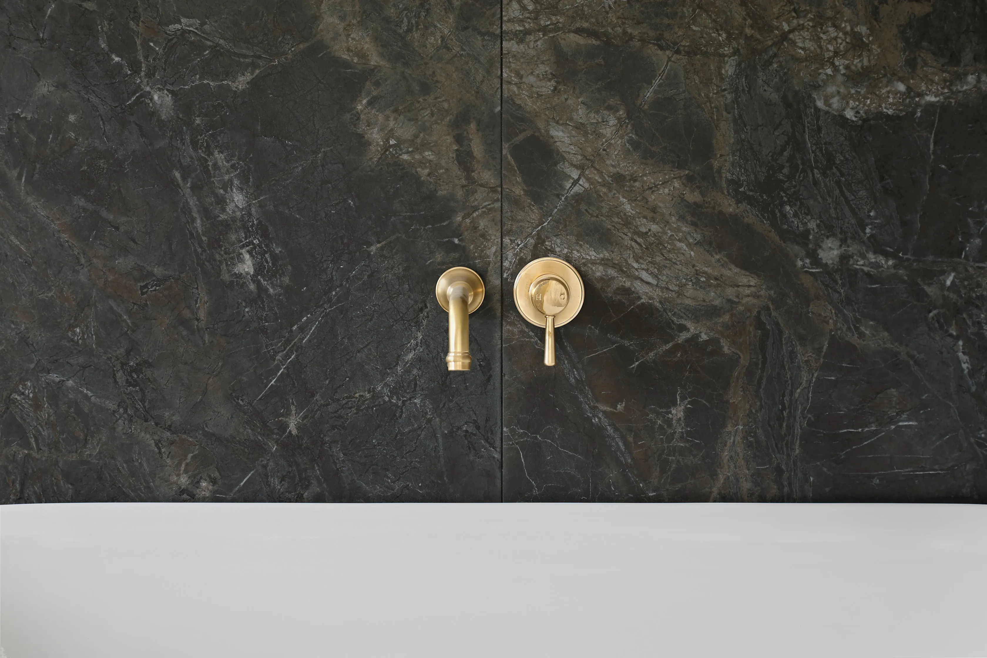 Main bathroom detail with a book-matched dark stone wall, aged-brass wall-mounted bath spout and mixer and a white freestanding bath