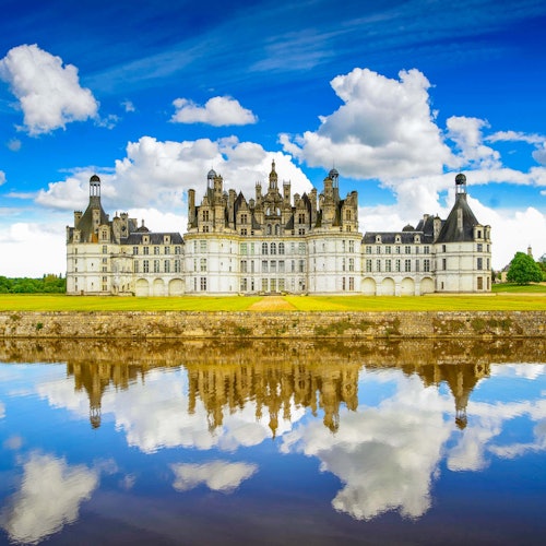 Un grand château avec des tours et tourelles ornées, entouré d'un fossé reflétant le ciel bleu et les nuages.