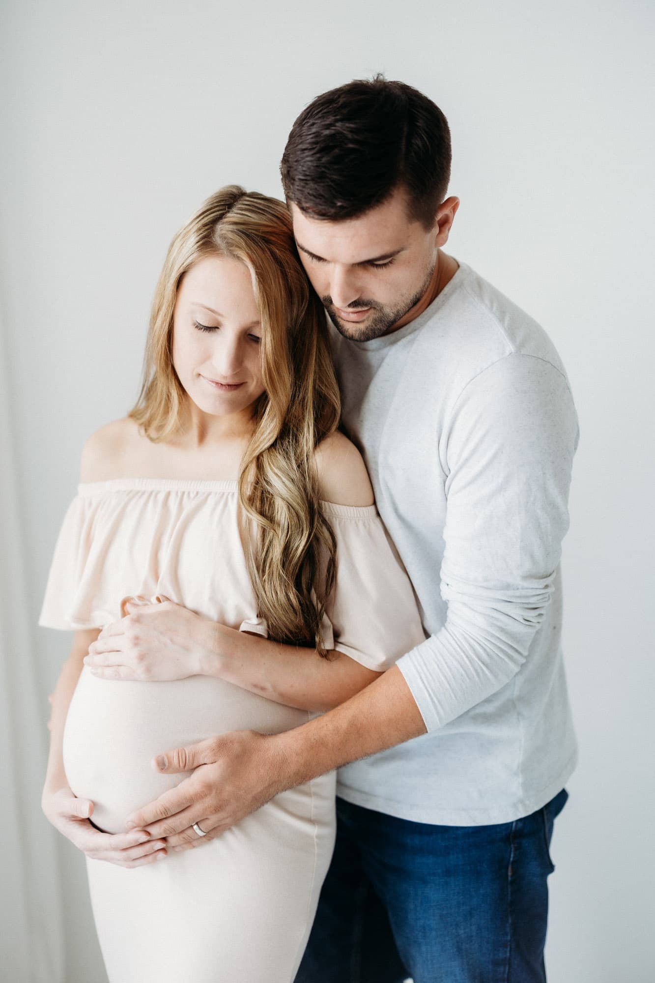 A pregnant mother gently cradles her belly while standing in a natural light studio in Shreveport, taken at Revelator Studio during a maternity session.