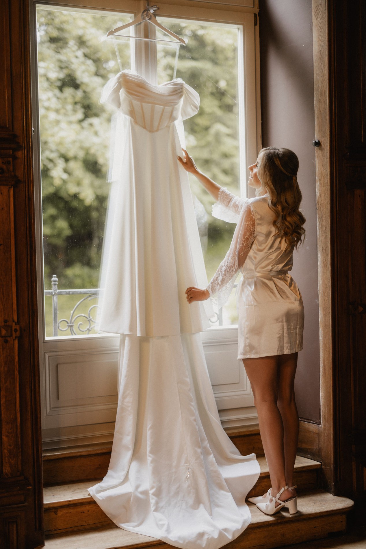 Braut am Fenster mit ihrem Hochzeitskleid während des Getting Ready