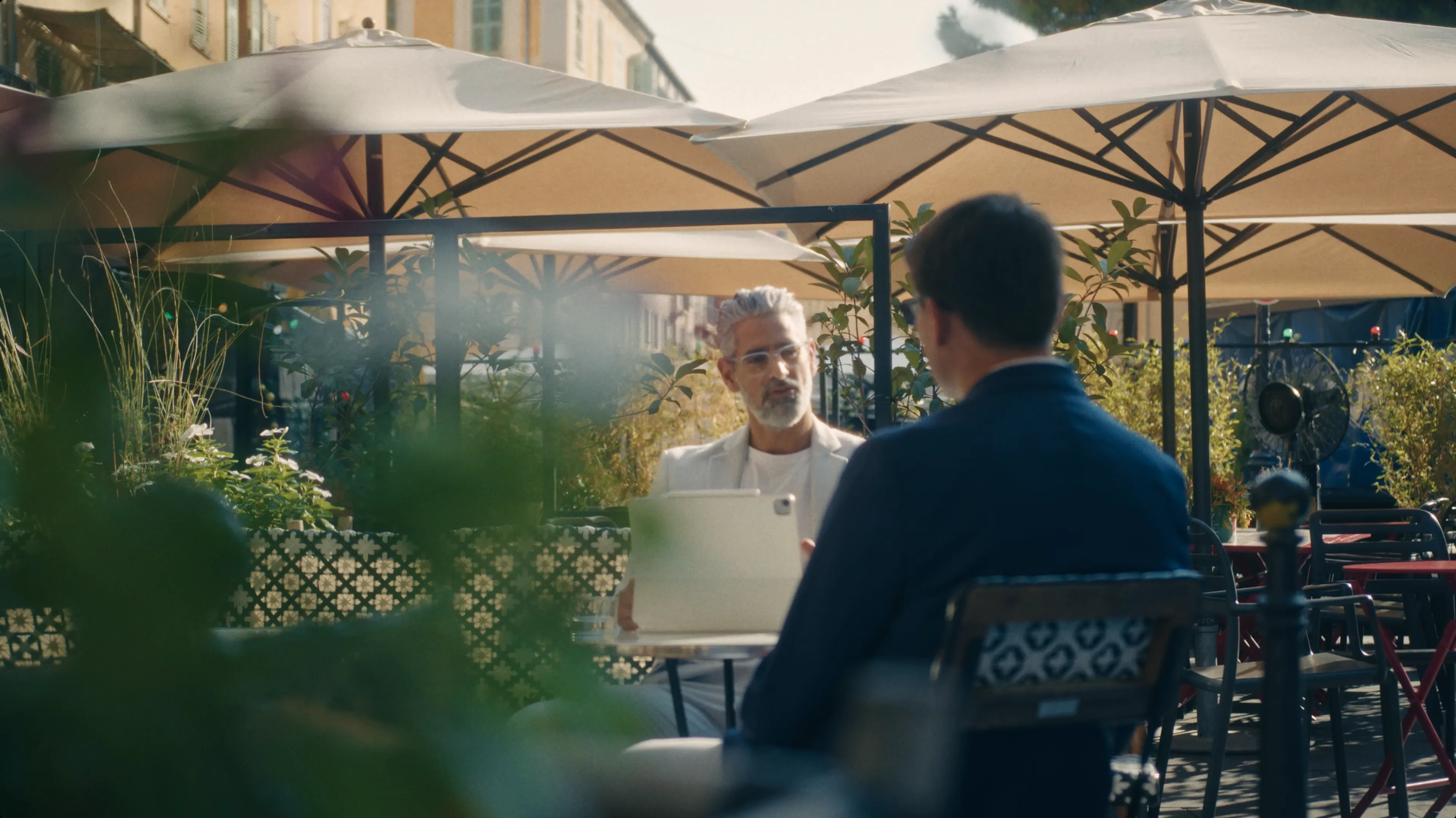 Two people sit at an outdoor café under large beige umbrellas, engaging in conversation amidst surrounding greenery and soft natural light.