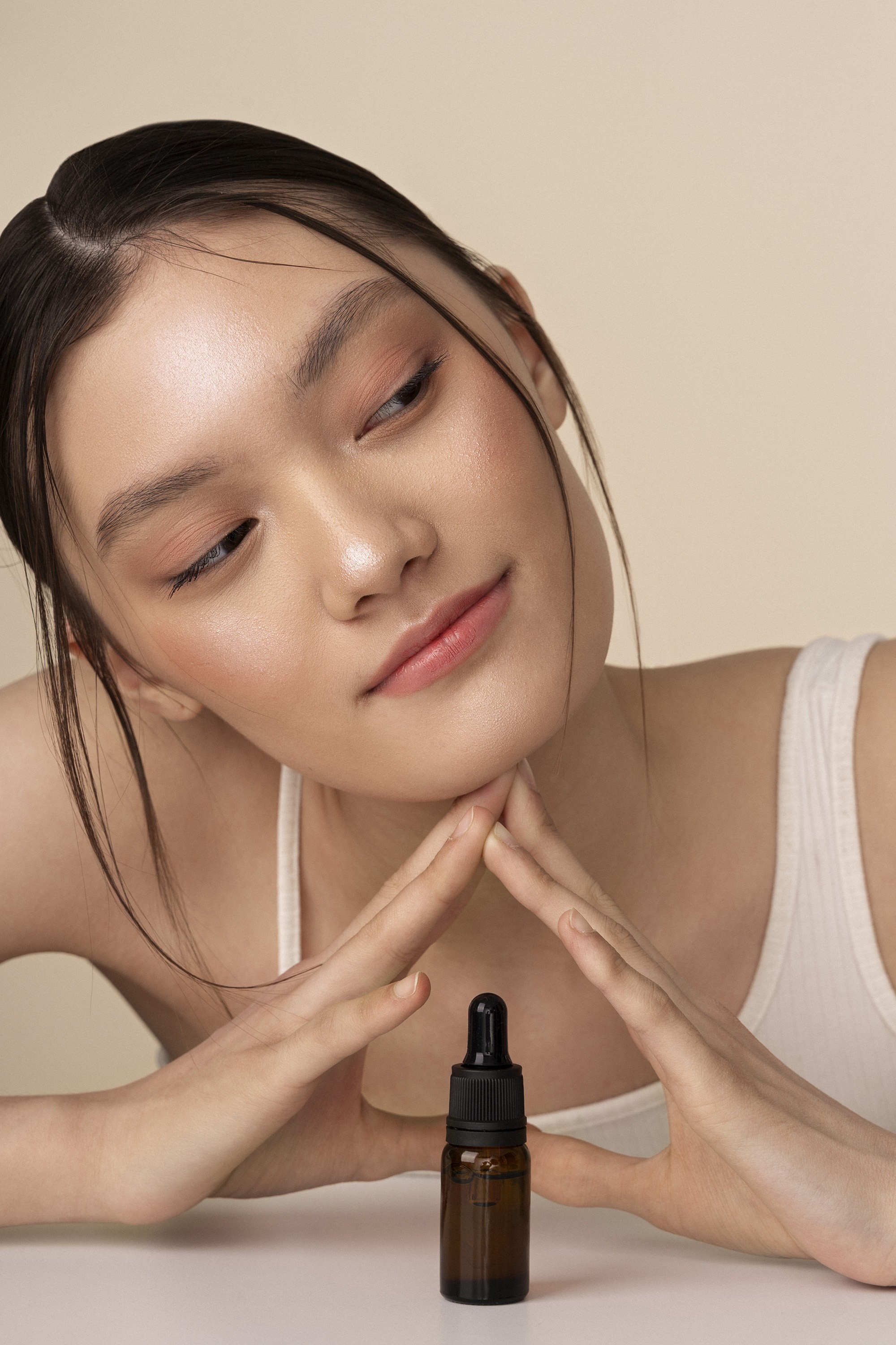 Young woman with a soft smile, resting her chin on her hands, gazing at a small serum bottle on a table