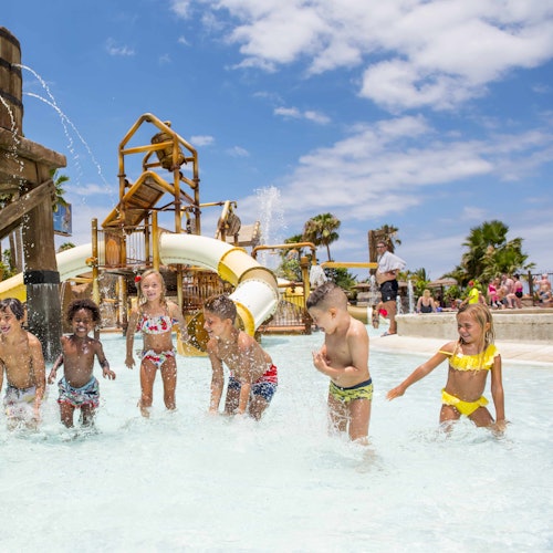 Children playing in a shallow water park pool with slides and water structures on a sunny day.