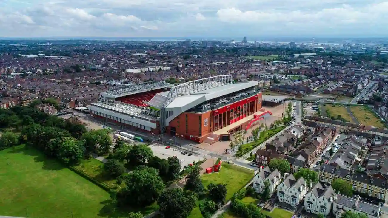 Aerial view of Anfield Stadium in Liverpool, surrounded by residential areas and green spaces. The stadium is the home of Liverpool FC, known for its intense atmosphere and the famous pressing game under Jürgen Klopp. Trees and parkland can be seen in the foreground, while the imposing stadium is at the centre of the picture.