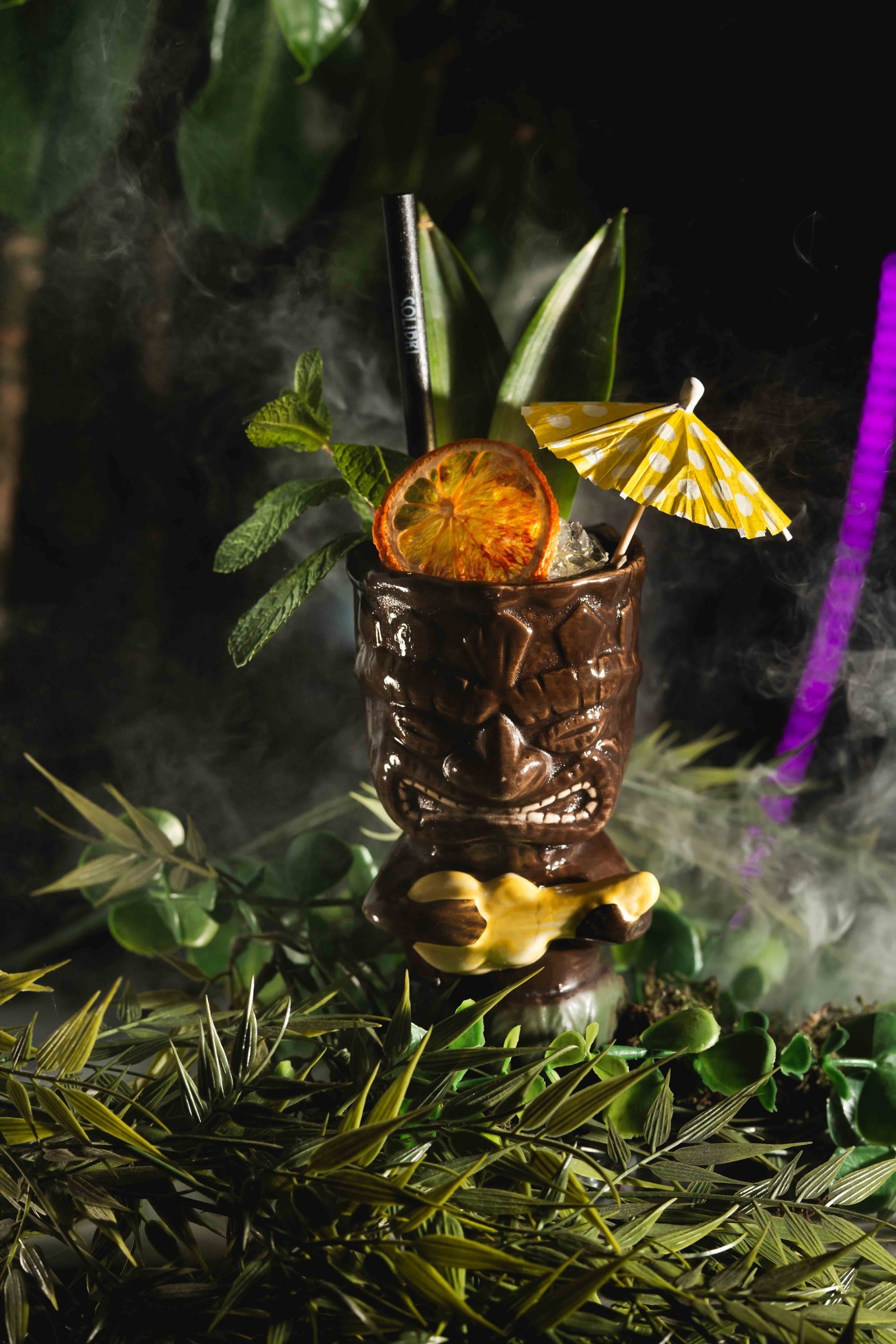 Cocktail servito in bicchiere con maschera hawaiana su foglie di palma e sfondo tropicale