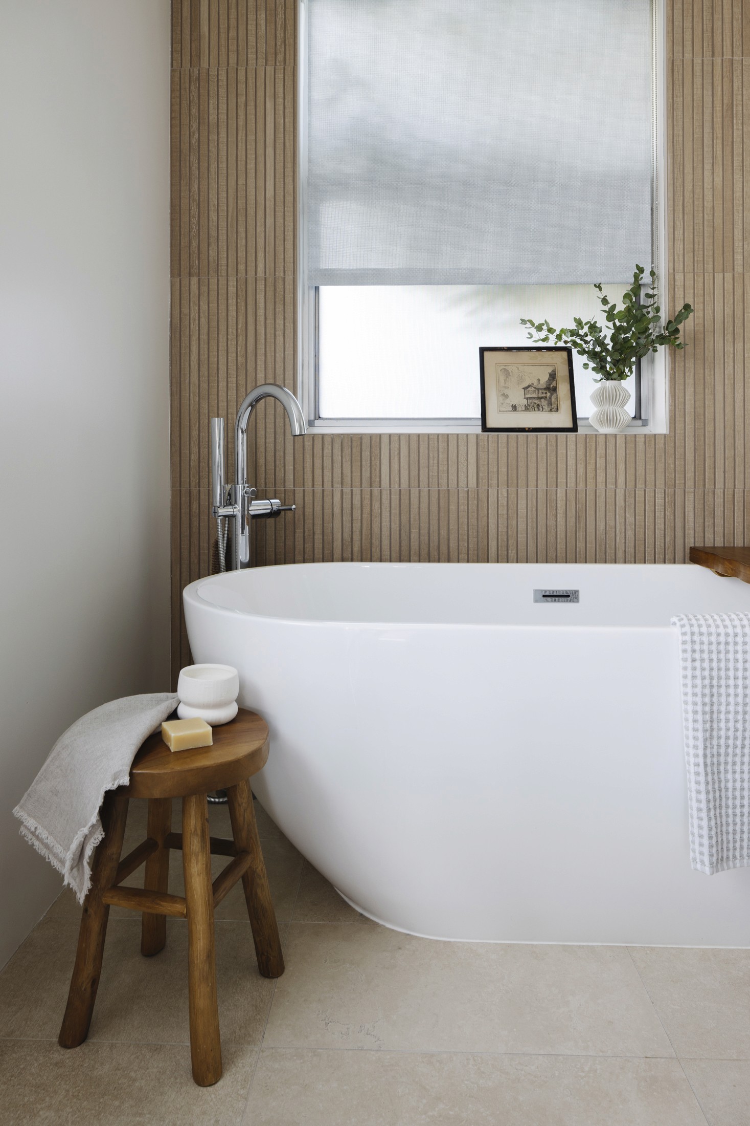 White freestanding bathtub with accent stool, window in background with vintage artwork and greenery