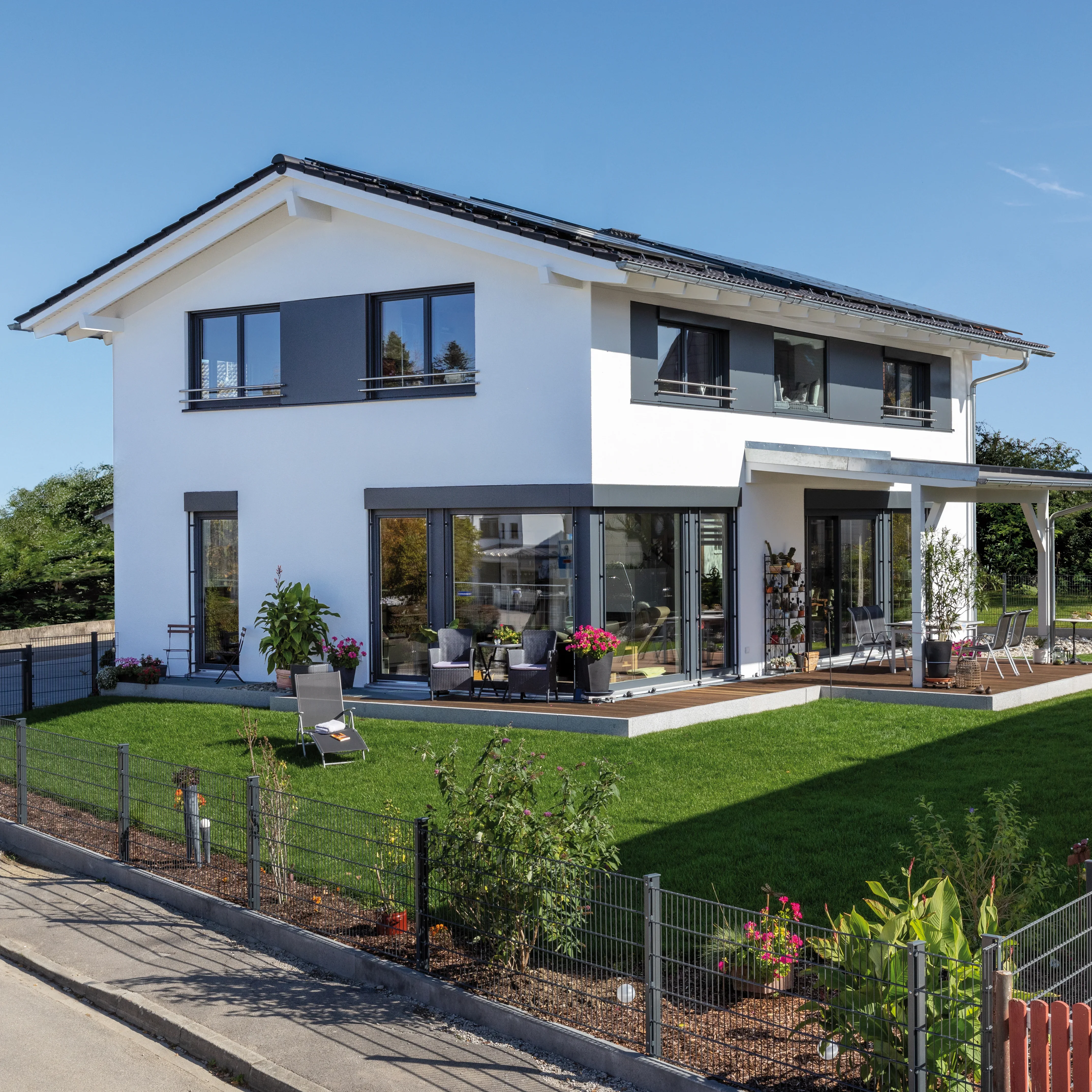 Modernes Einfamilienhaus mit weißer Putzfassade, dunklen Fensterrahmen und Solaranlage auf dem Dach, mit Terrasse und Garten.