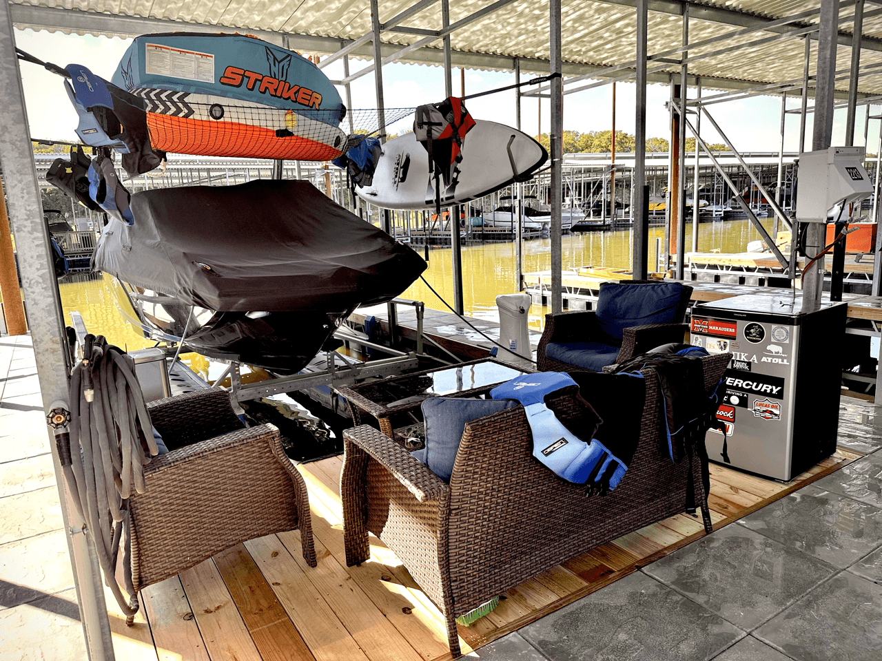 The image shows a boat dock setup featuring a covered boat lift with an orange and turquoise "Striker" boat, accompanied by kayaks on a storage rack, situated under a shaded, metal canopy with nearby seating including a wicker armchair, table, and life jacket, all positioned on a wooden deck by a marina dockside.