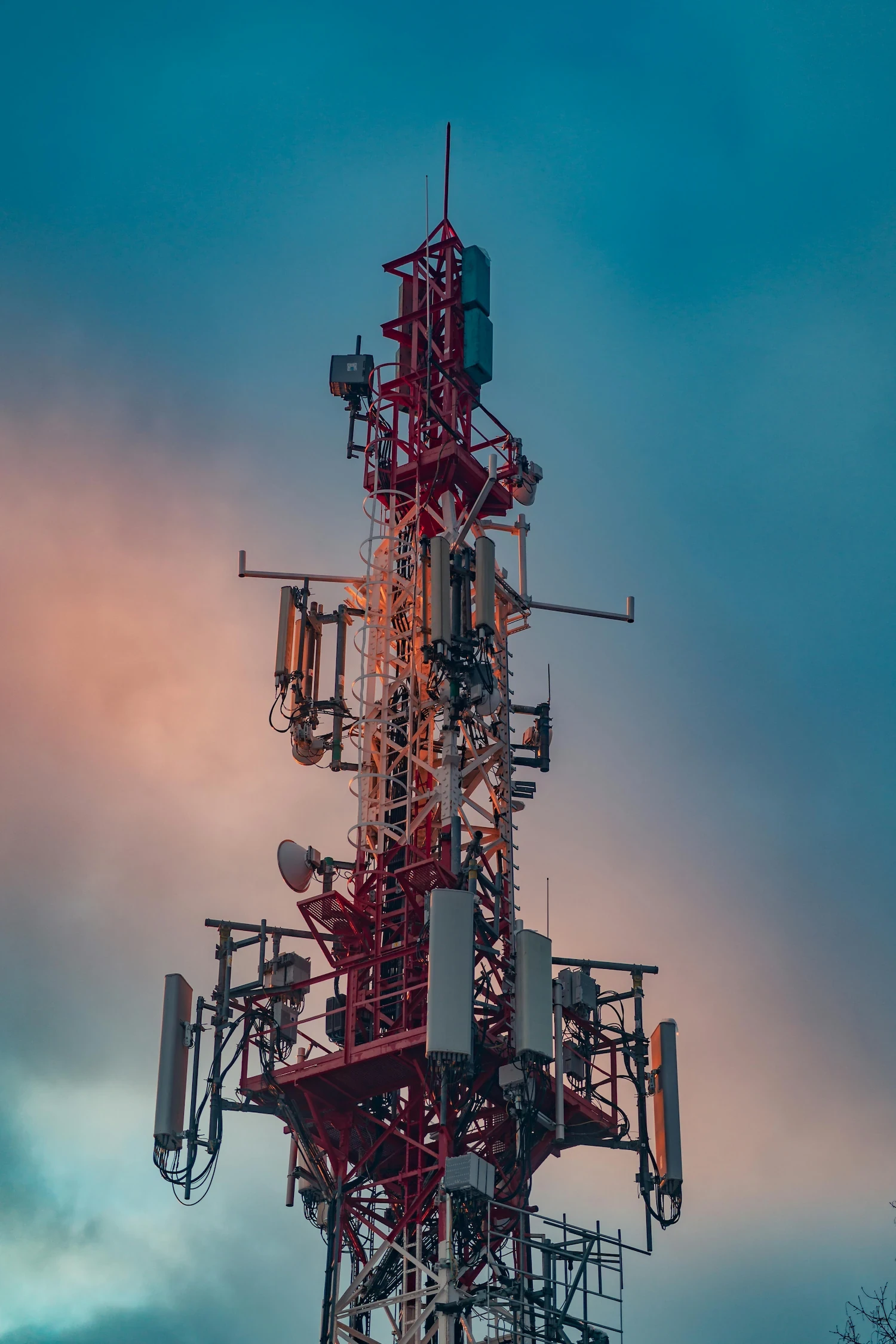 Tall red and white lattice telecoms tower equipped with cellular antennas and microwave dishes against a dusk sky
