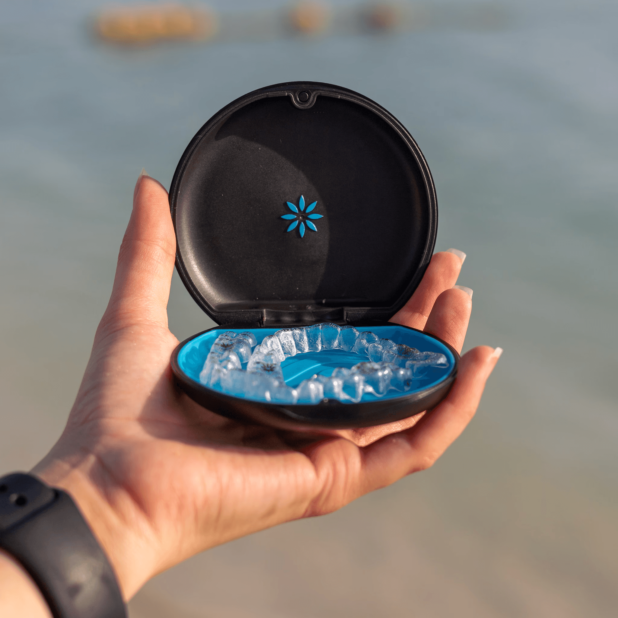 يد تمسك علبة تقويم أسنان سوداء ببطانة زرقاء تحتوي على قوالب انفزلاين الشفافة، مع خلفية هادئة لساحل البحر توحي بالراحة والحرية التي يوفرها تقويم الأسنان.