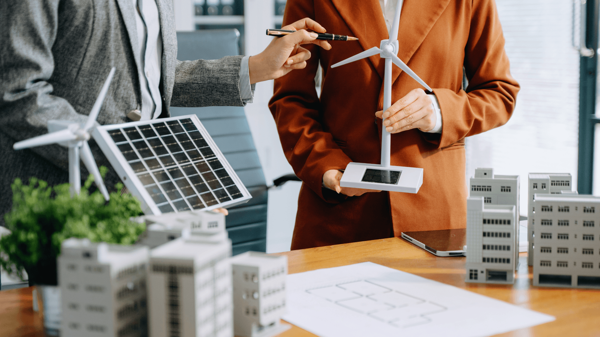 A close-up photo of two business professionals meeting at a table to discuss renewable energy projects. The table features models of wind turbines and solar panels, along with scale models of buildings and a technical drawing of a project plan.