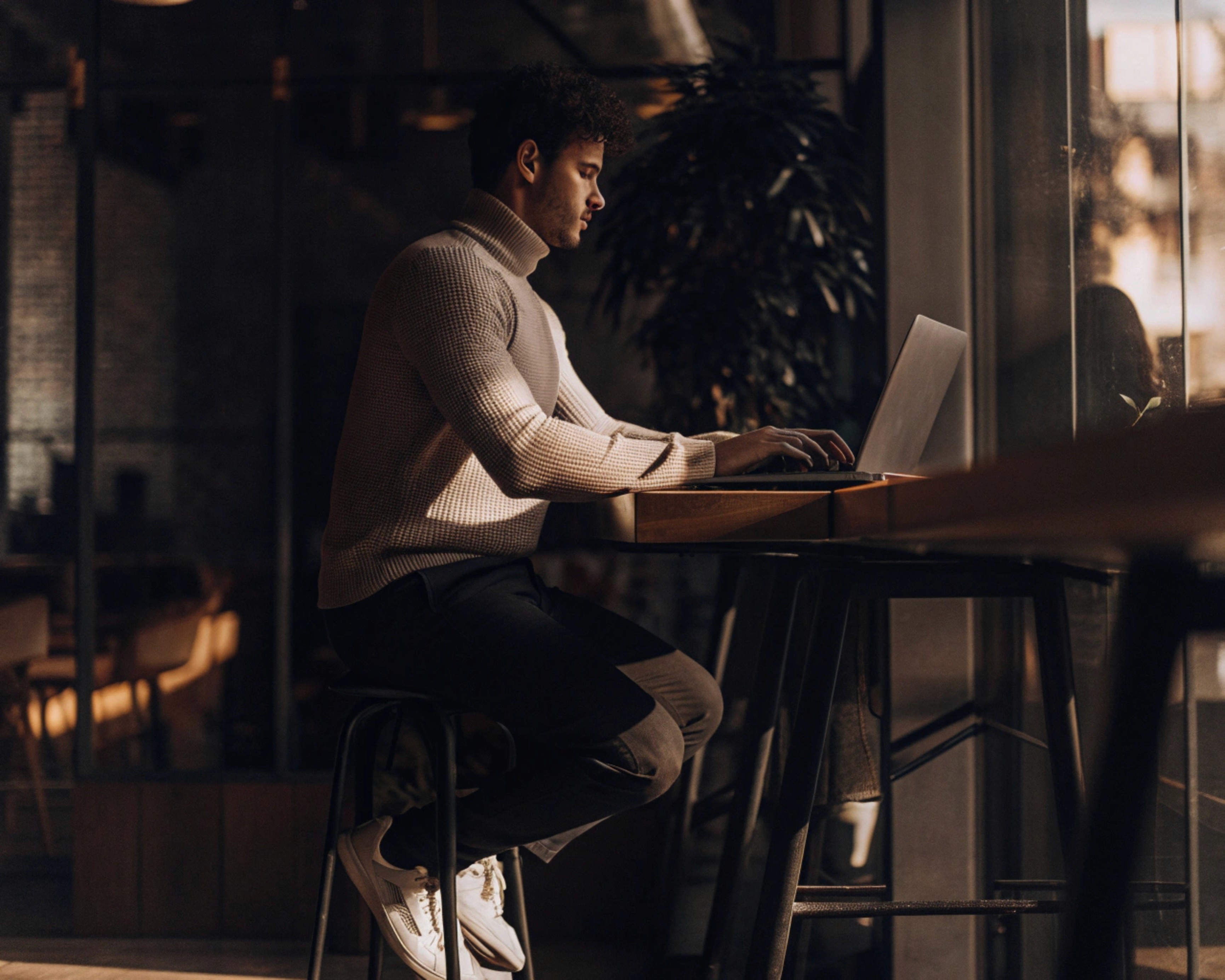 Man in turtleneck sweater using a laptop at a table, working or studying. Indoors, natural light.