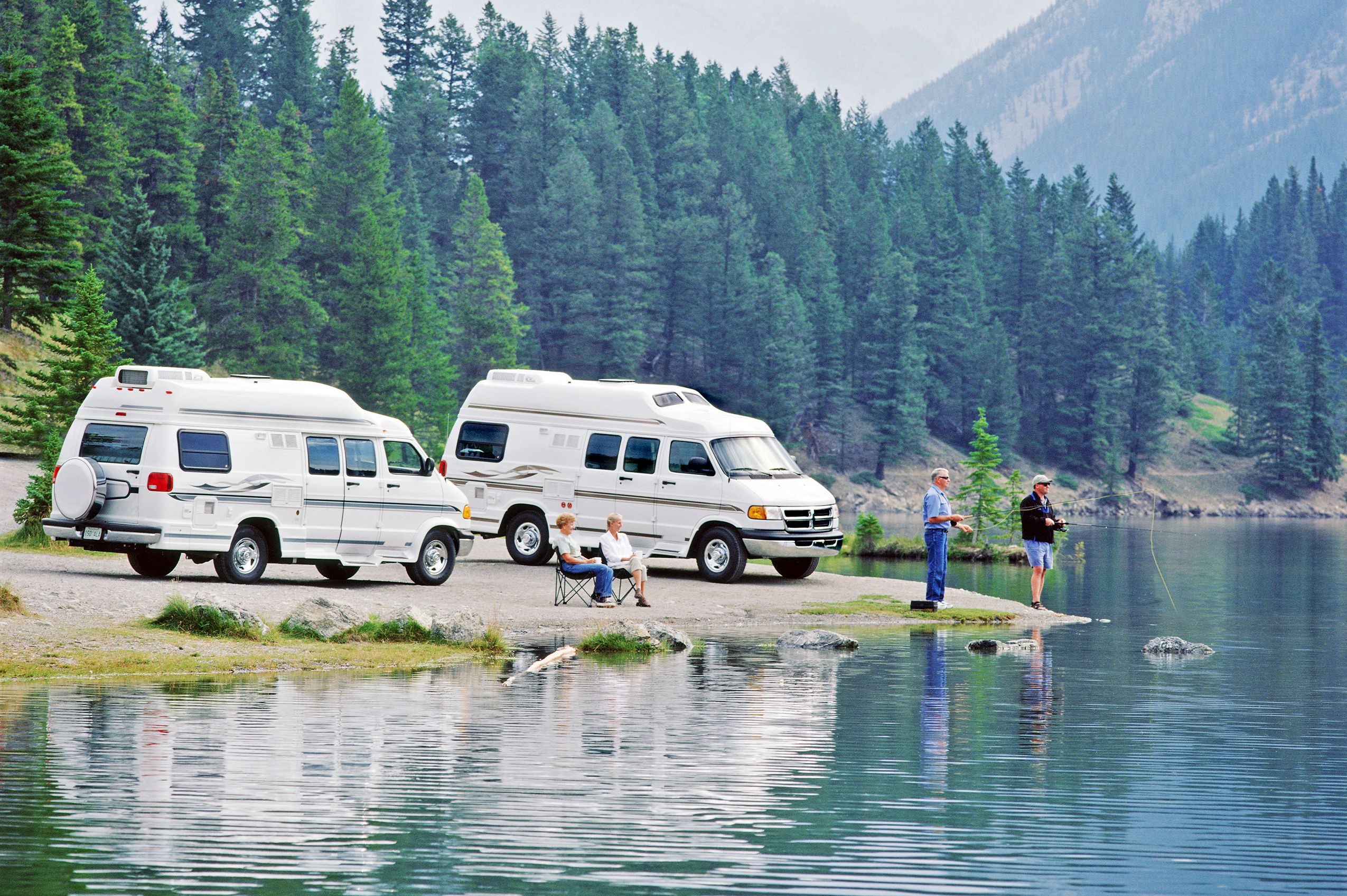 Two white camper vans by lake with people fishing