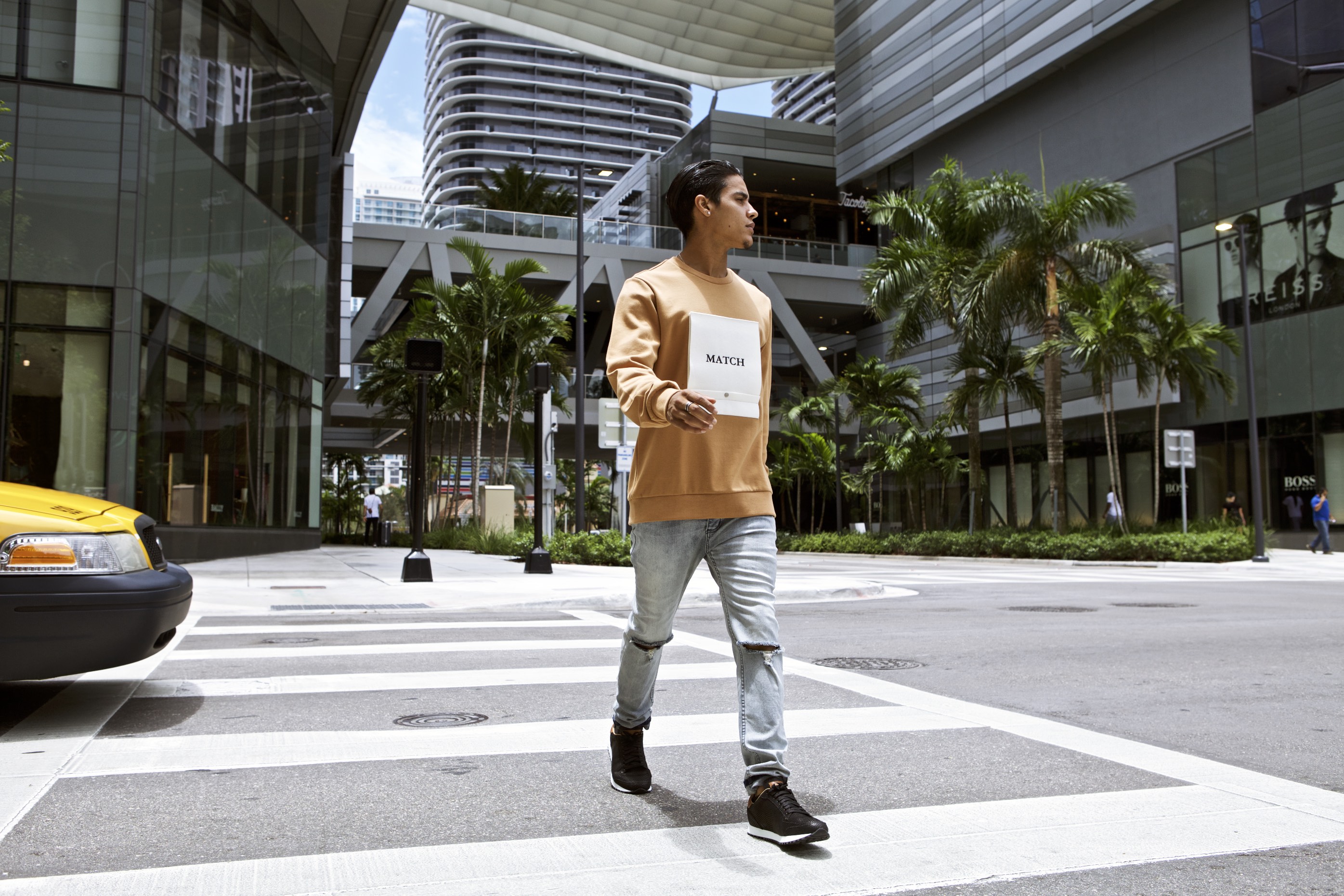 Model in a tan sweatshirt and ripped light-wash jeans crossing a Miami street, palm trees and modern buildings in the background.