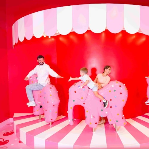 三个人正在一个色彩鲜艳、以马戏团为主题的环境中骑着粉色波点的大象座椅，周围是红色和粉色的装饰。