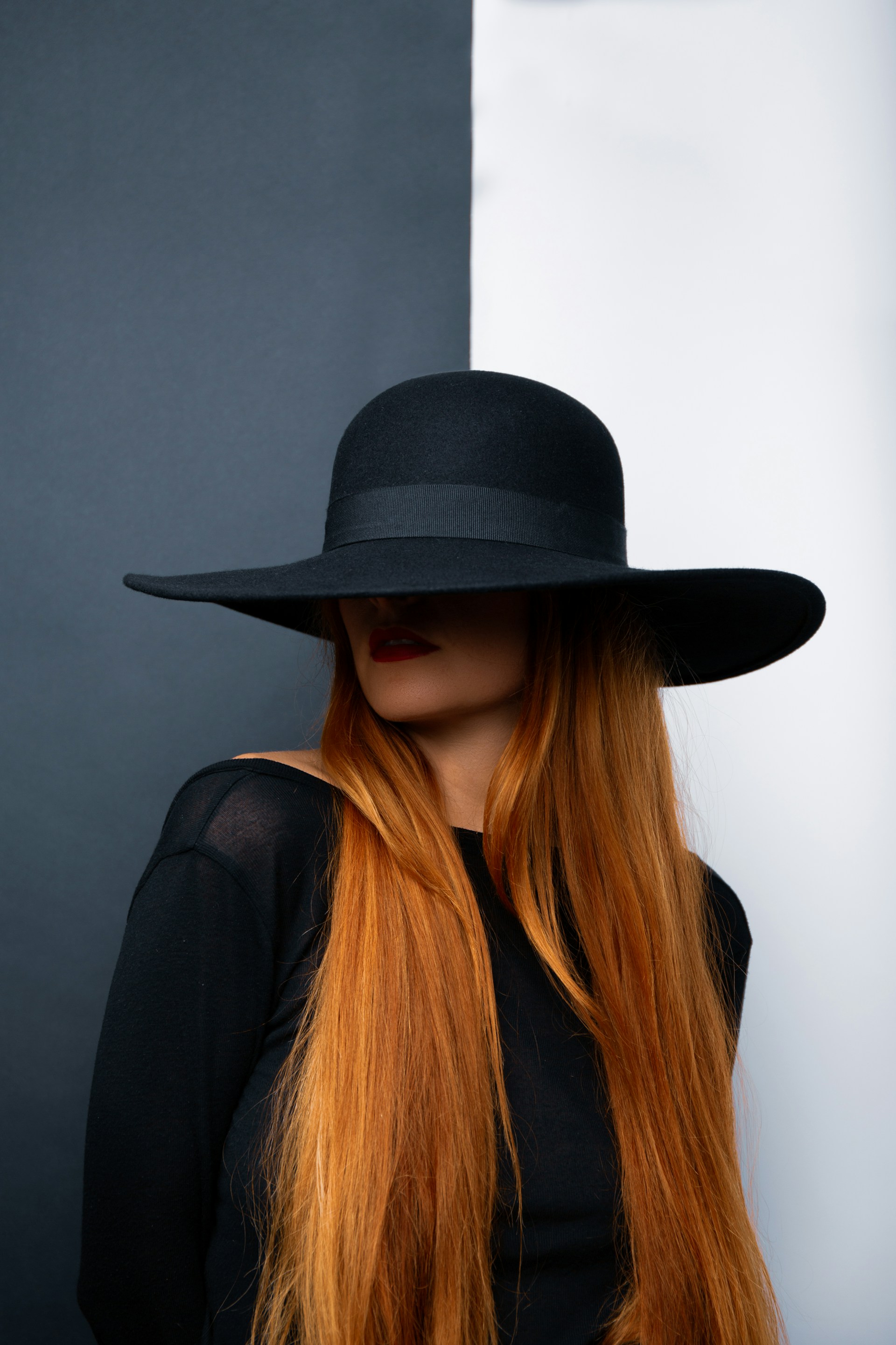 A woman with very long, straight ginger hair wearing a wide-brimmed black hat that shadows her eyes. She is wearing a black top and red lipstick, posed against a split black and white background.