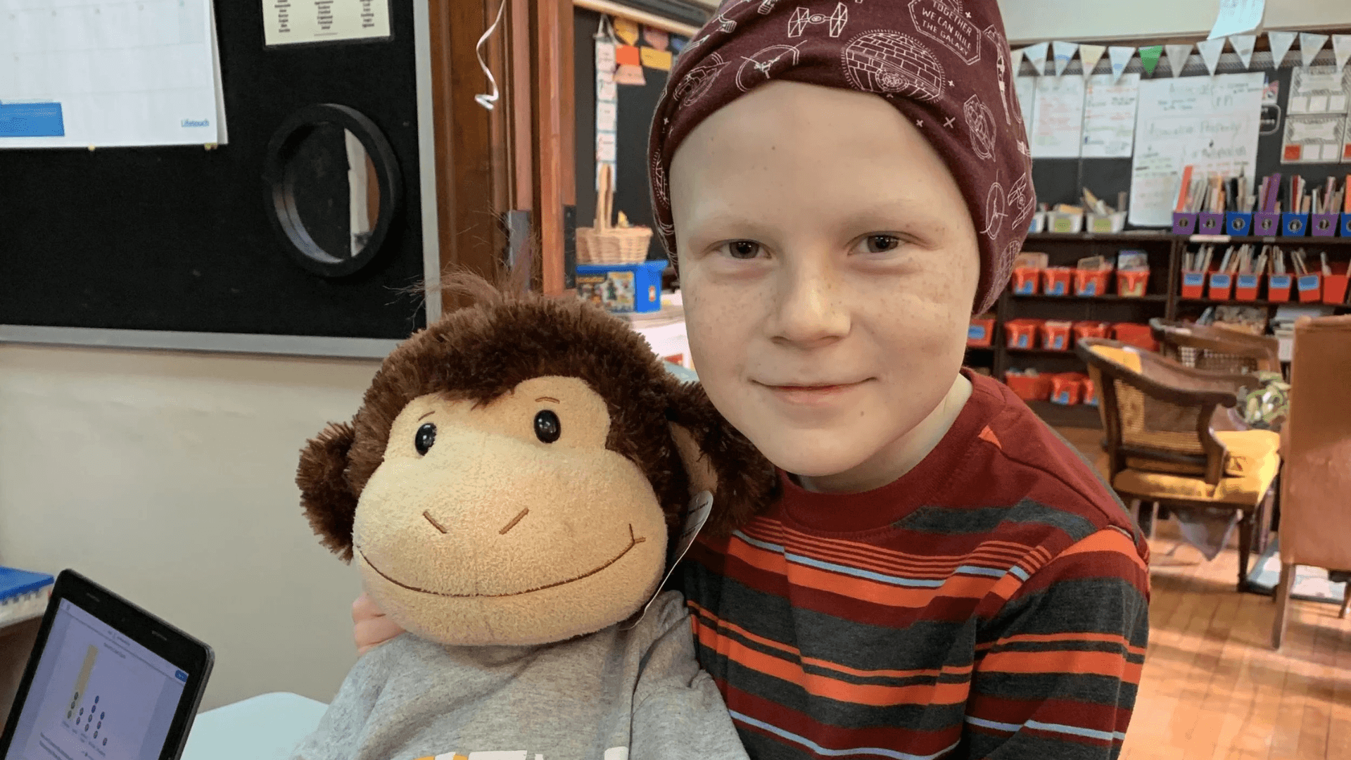 Smiling child wearing a patterned headscarf and striped shirt holding a stuffed monkey in a classroom setting.