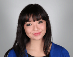 The image shows a young woman with long, dark hair styled with bangs that fall across her forehead. She is wearing a blue cardigan over a dark-colored top. The background is a plain, light gray or off-white color. Her gaze is directed straight at the camera, and she has a neutral facial expression with light makeup that includes eyeshadow and possibly lip gloss or lipstick.