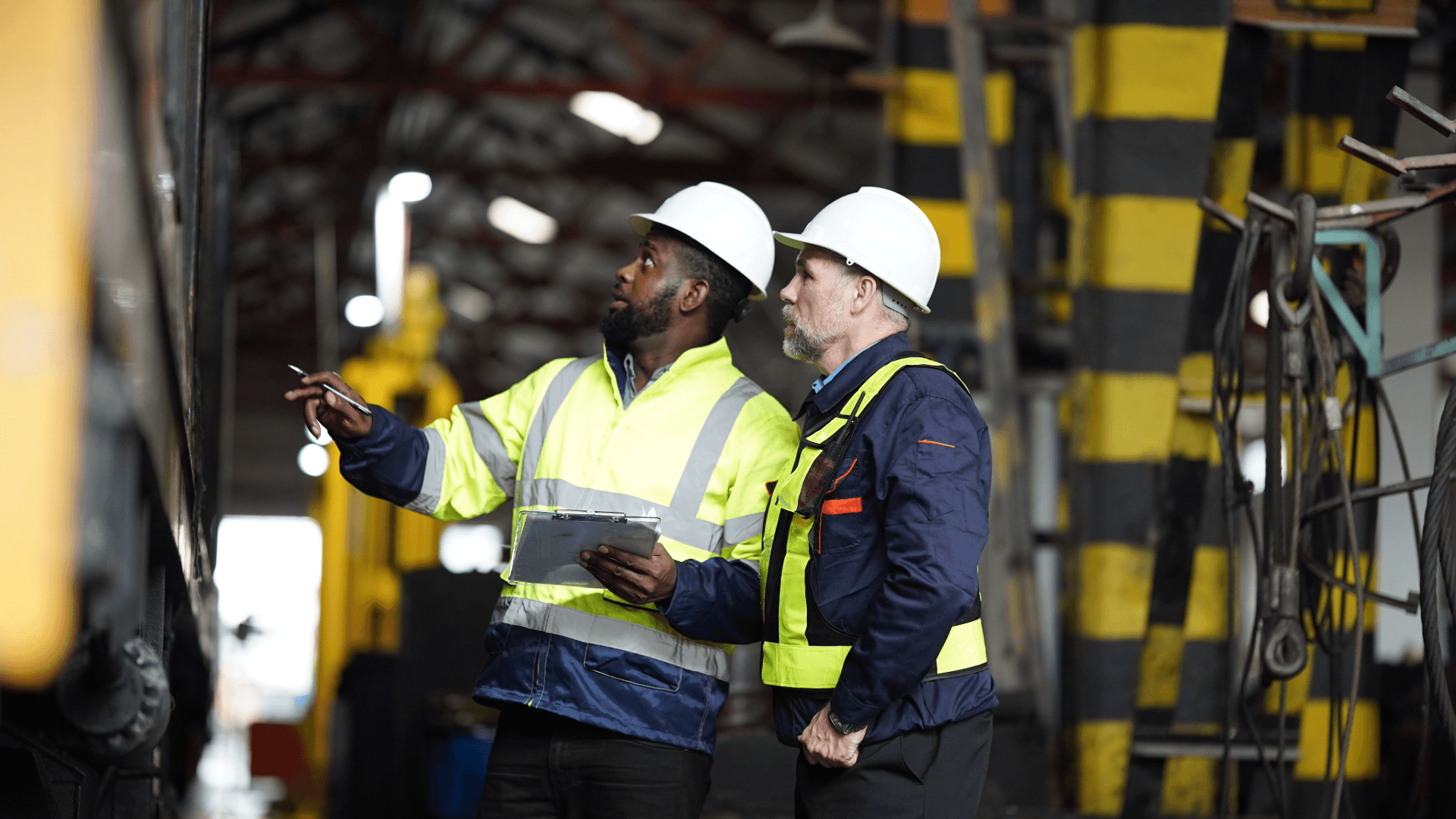 Facilities management engineers inspecting industrial equipment during maintenance inside a workshop, wearing high-visibility PPE and safety helmets.