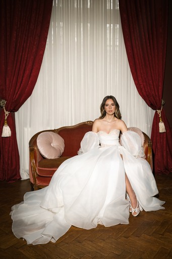 Mireasă elegantă în rochie de bal albă, așezată pe canapea vintage cu perdele roșii — ședință foto nuntă București