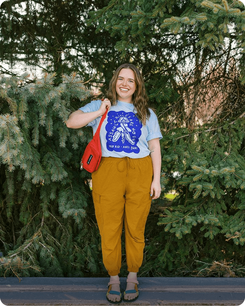 Woman smiling, wearing a blue t-shirt with a design, mustard yellow pants, and a red bag. Standing in front of evergreen trees.
