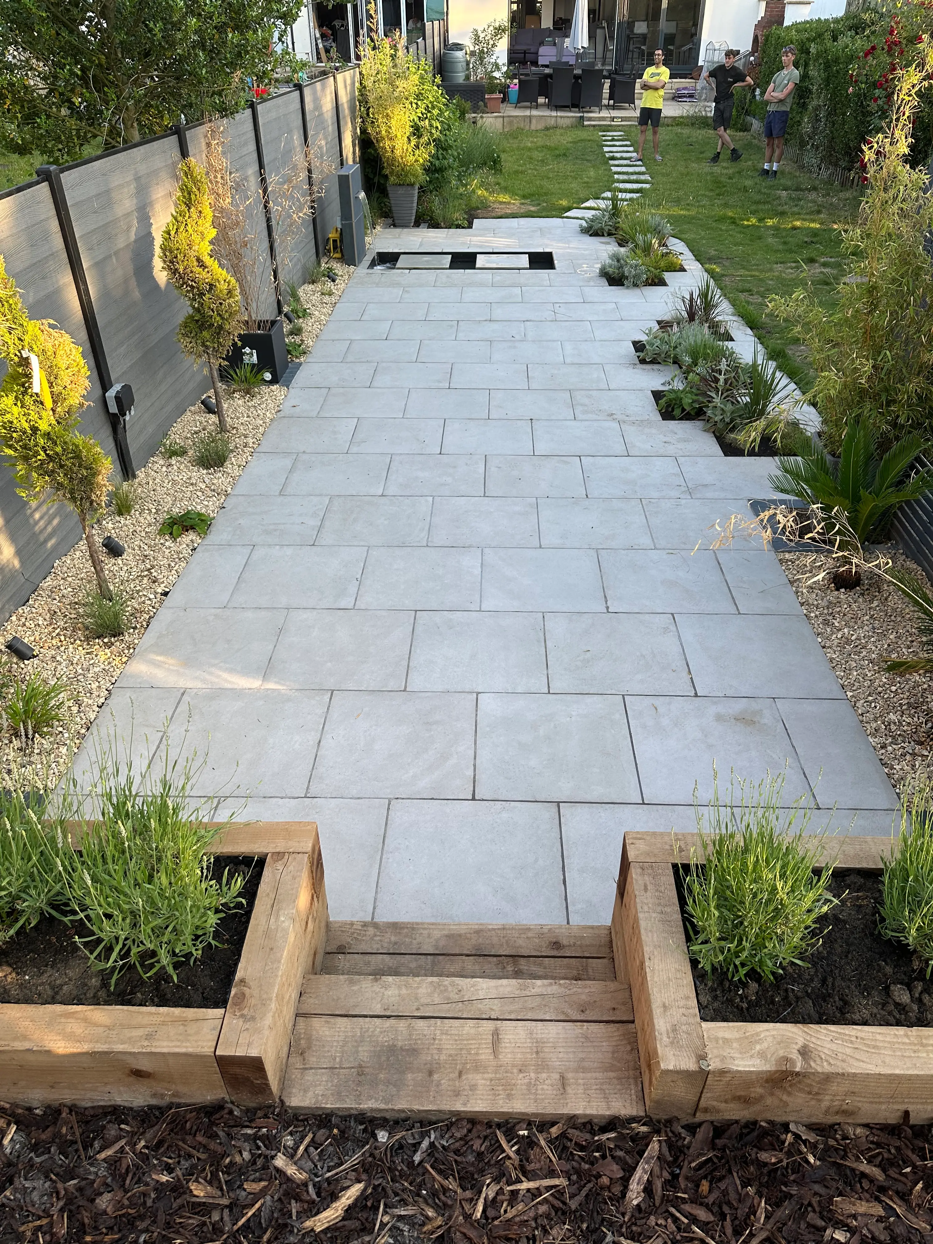 A stone pathway leads through a garden, flanked by plants and yellow flowers, with brick borders on either side.