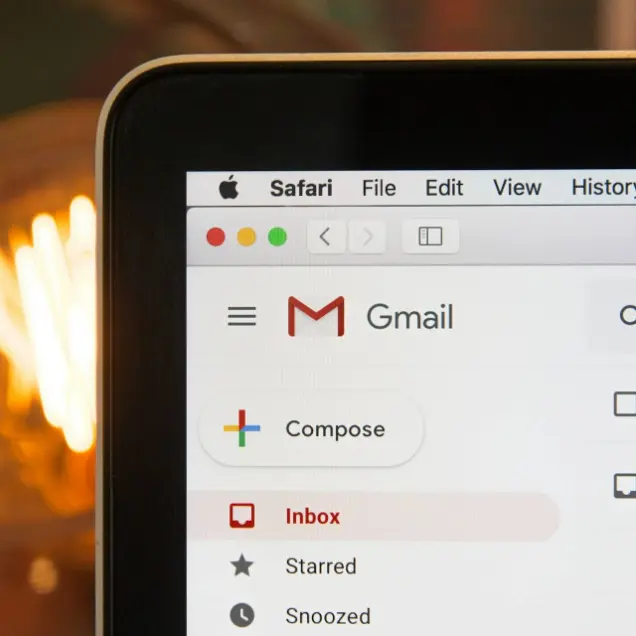 Close-up of a laptop screen showing the Gmail interface in Safari browser. Visible are the Compose button, Inbox, and menu icons. Warm lighting in the background.