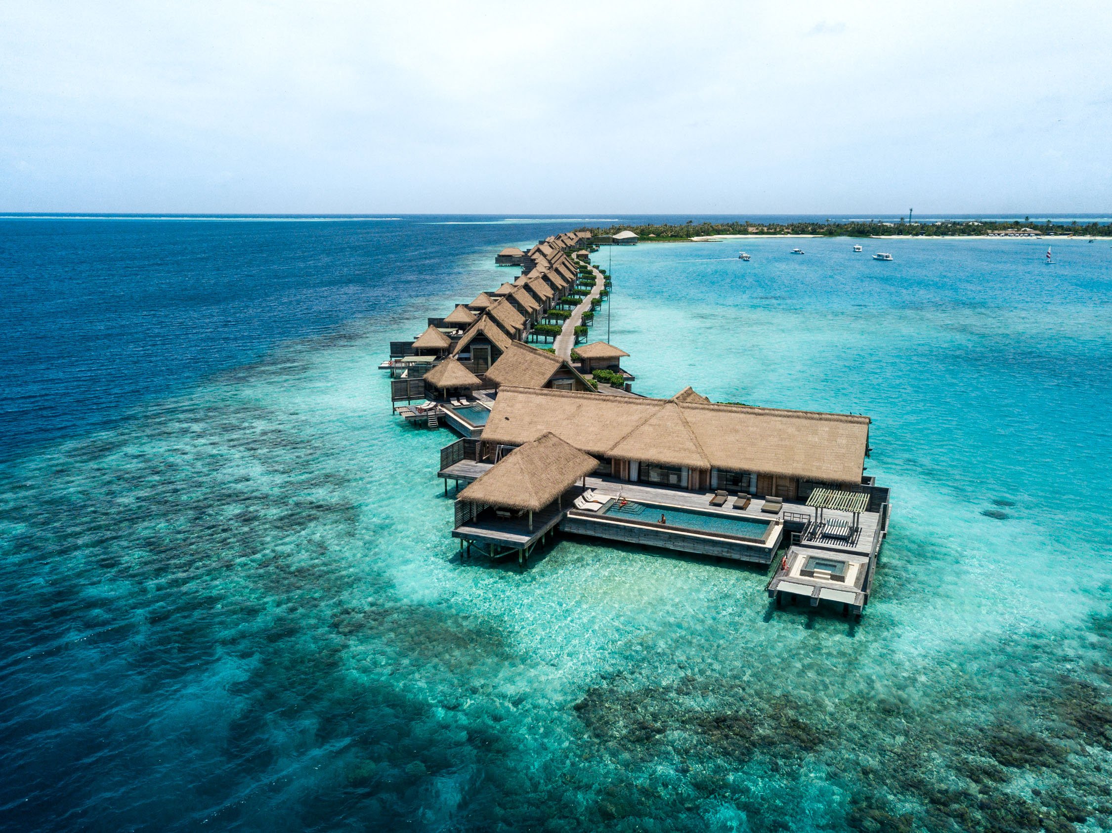 Overwater bungalows with thatched roofs line a turquoise lagoon in a tropical ocean under a clear sky.