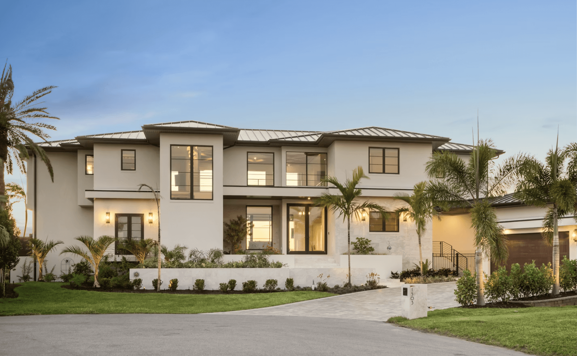 Luxury modern beige stucco mansion with large glass windows, palm trees lining the driveway and manicured lawn under clear blue sky.
