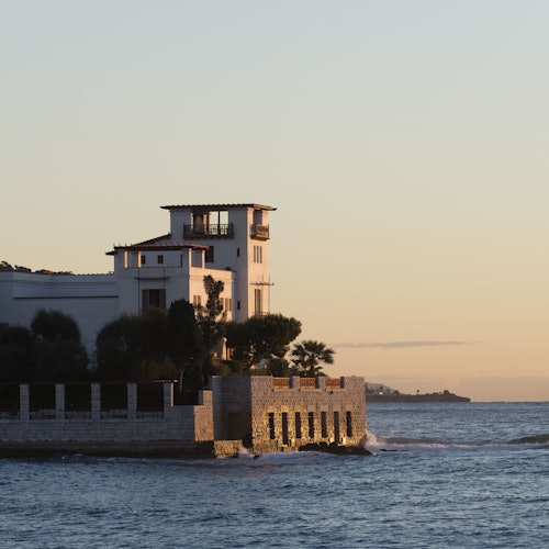 一座有塔樓結構的海邊別墅矗立在海岸線上，夕陽下四周被水環抱。