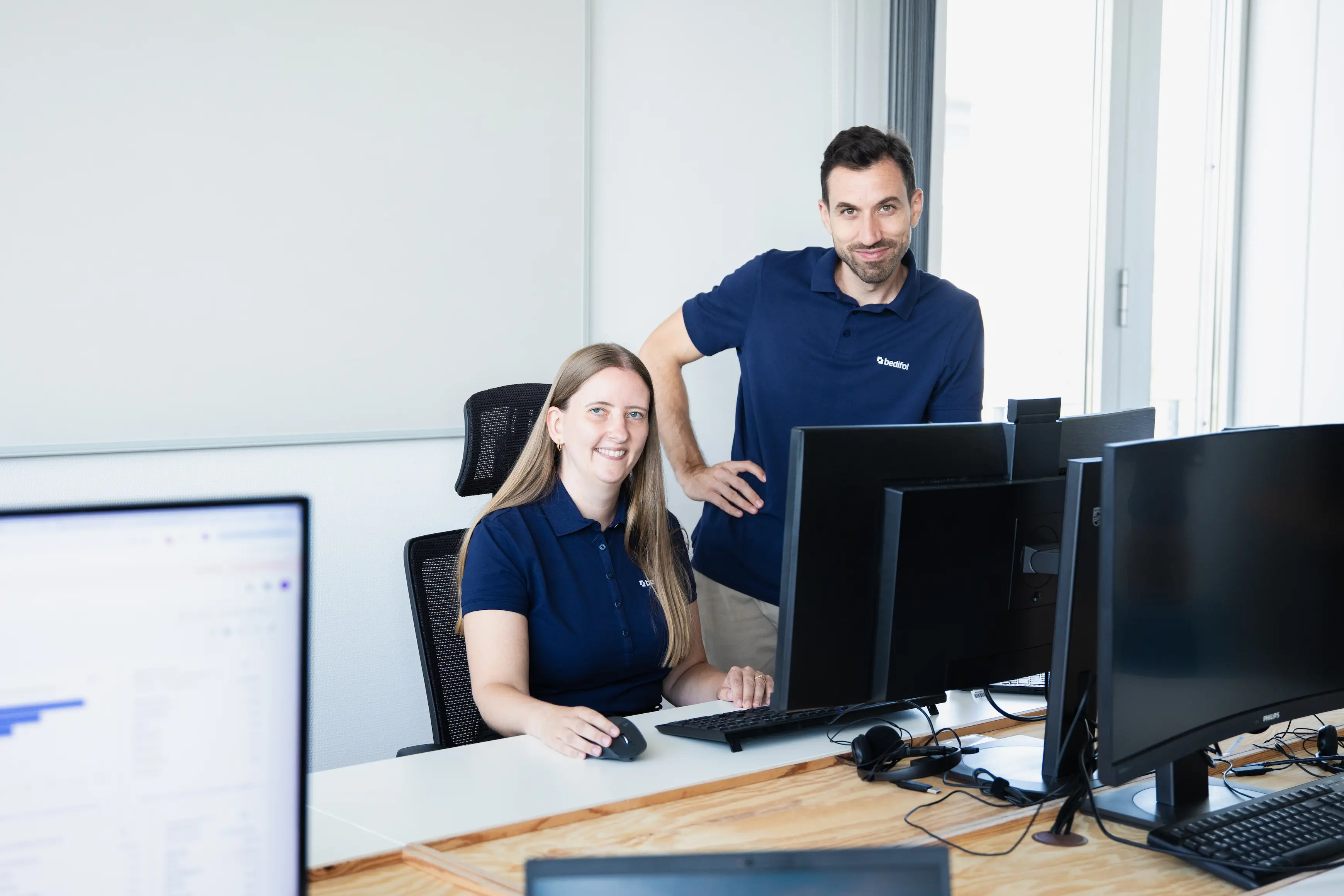 Zwei Mitarbeitende in Bedifol-Poloshirts an mehreren Bildschirmen im Büro, eine Person sitzt, eine steht.