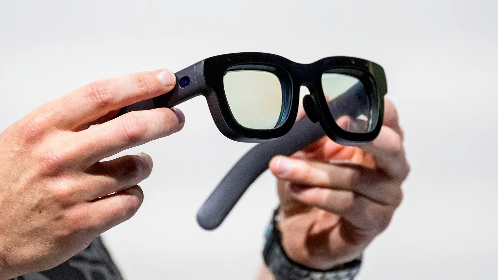 Hands holding a pair of black smart glasses with tinted AR lenses, camera sensor visible on the left temple