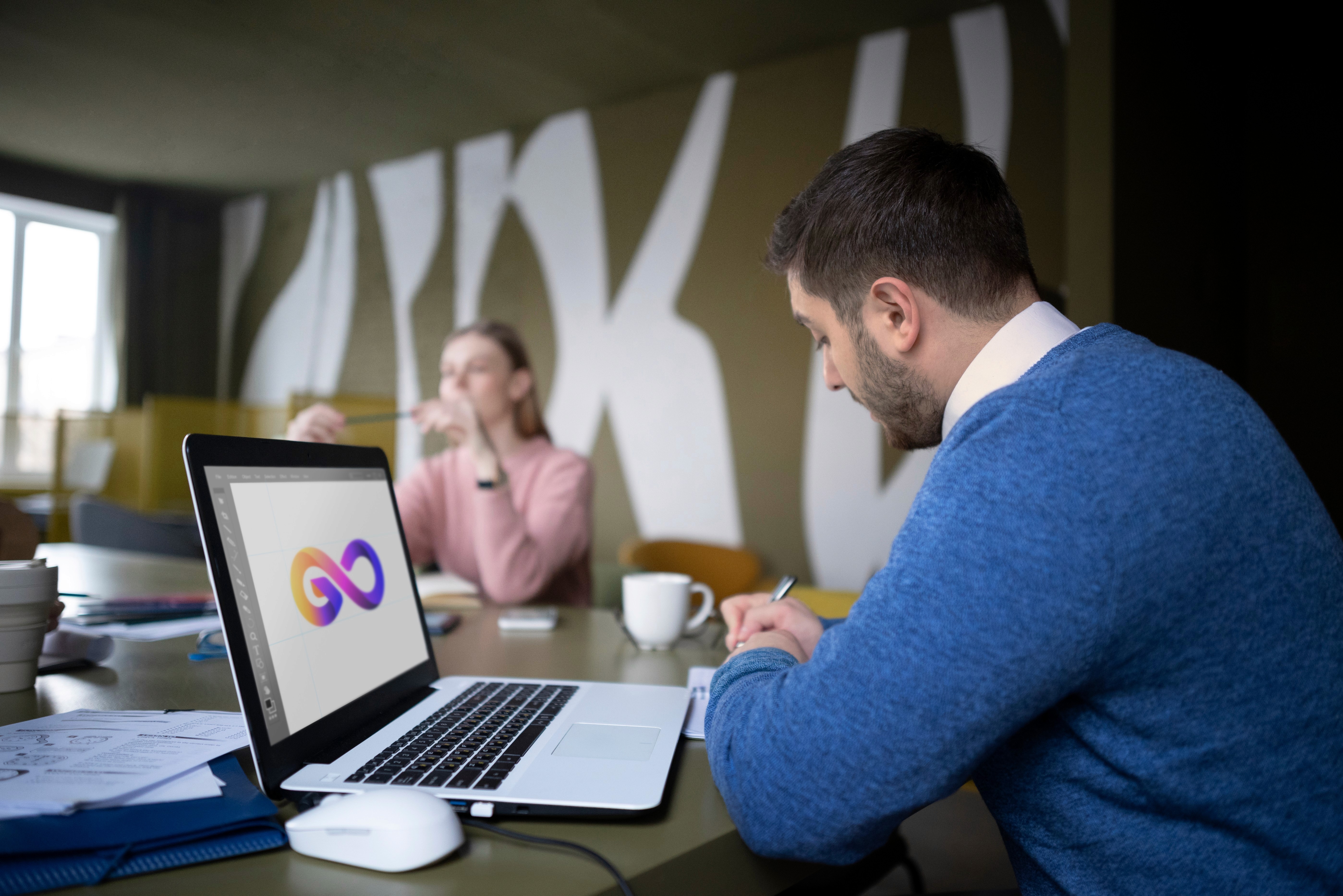 AWS DevOps professional designs infinity logo on laptop while collaborating with colleague in modern office space