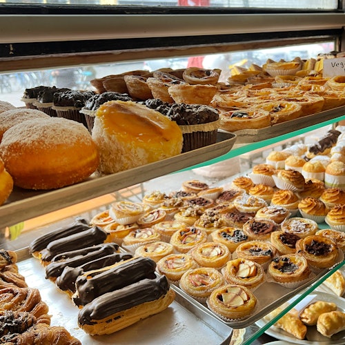 A variety of pastries including eclairs, tarts, and donuts displayed on metal and glass shelves in a bakery.