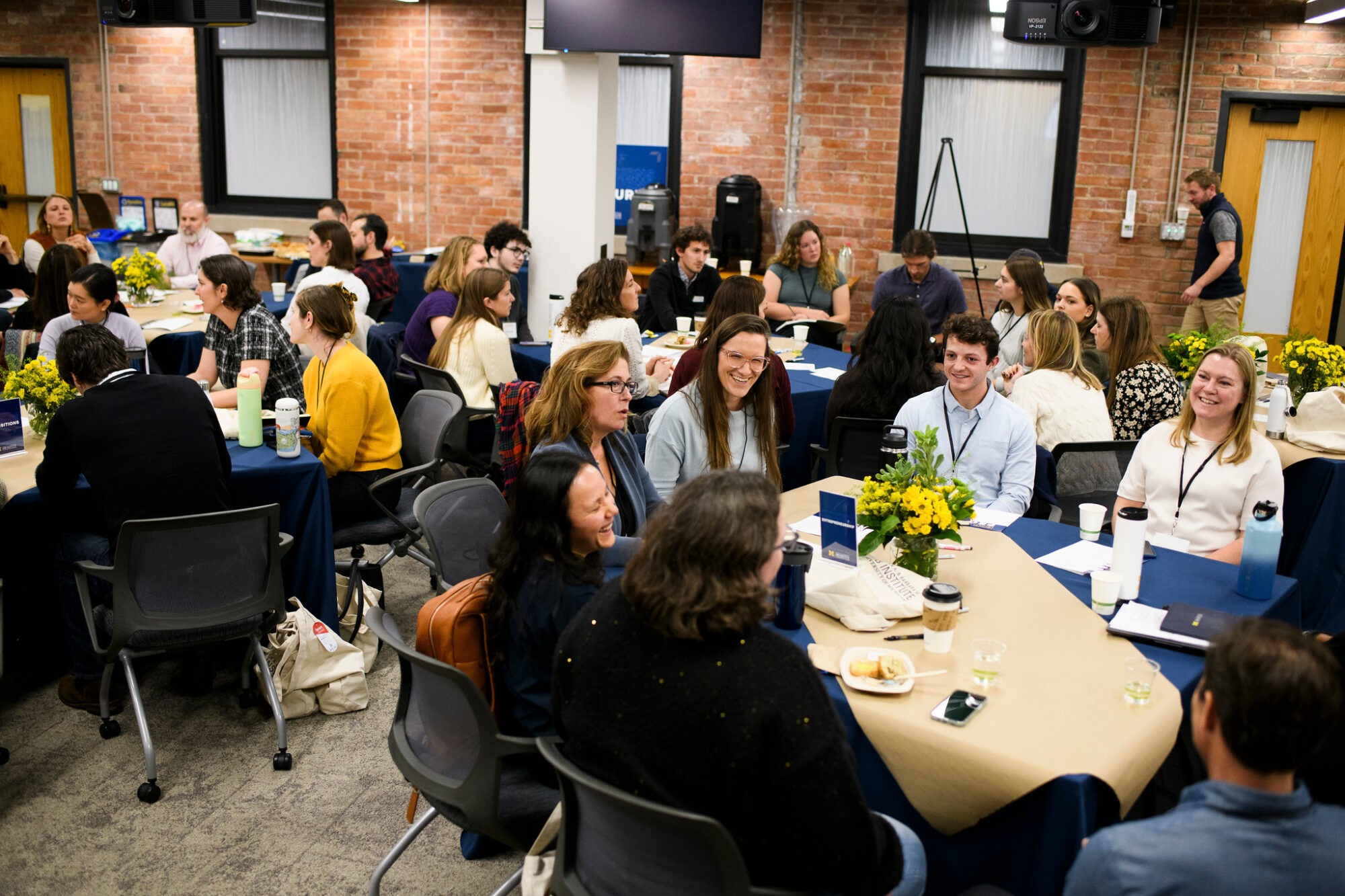 A group of people gathered around tables in a bright, informal setting, engaged in conversation and enjoying refreshments.